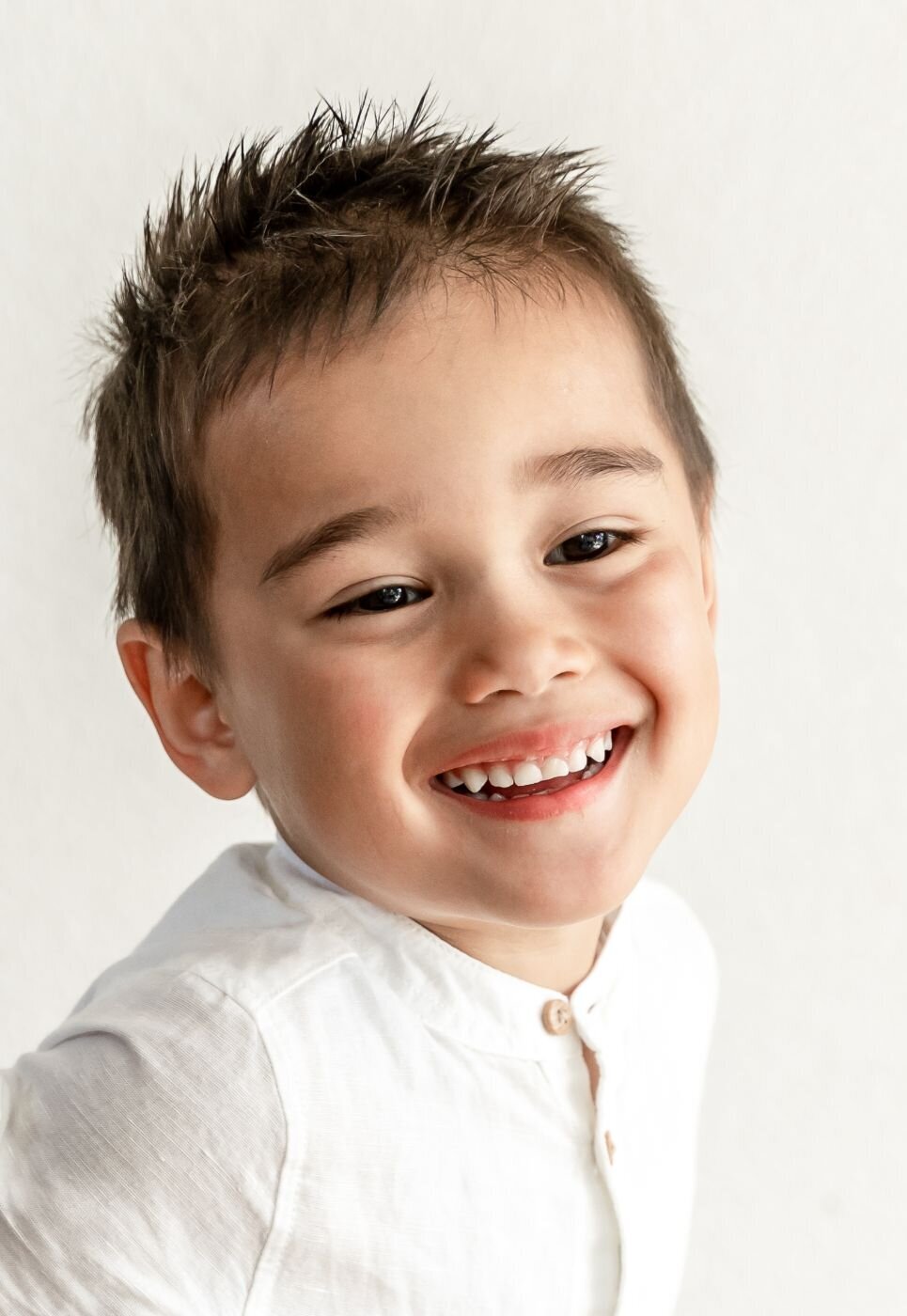  A close-up color portrait of a young boy with a big, happy smile. He is looking directly at the camera. He has dark hair styled in a spiky faux hawk and is wearing a white shirt with a button collar. The background is a simple, soft white.