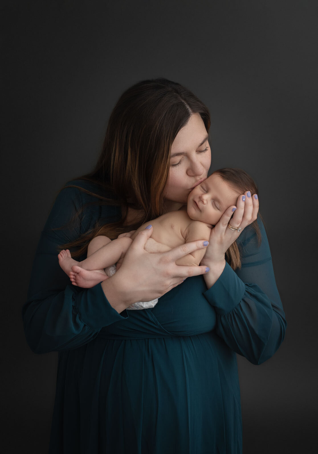 mom kissing her naked newborn on the cheek while wearing teal dress on gray background