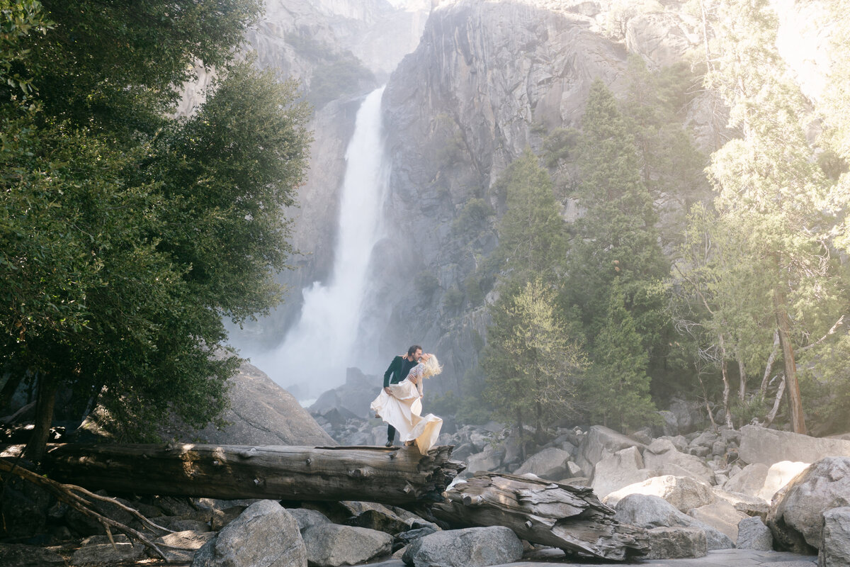 Bride and groom at Yosemite waterfall during golden hour the groom dipping the bride and the wind blowing the Claire Pettibone dress and her hair in a dramatic way