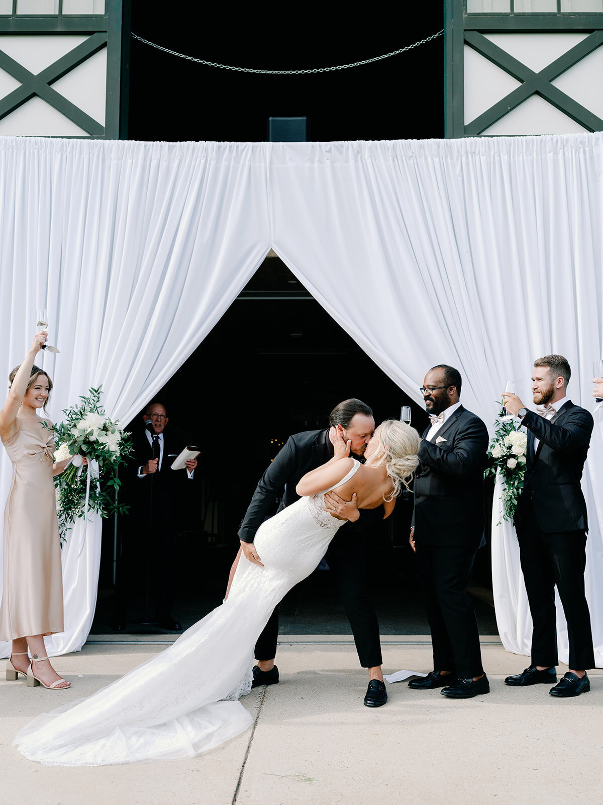 Bride and groom share their first kiss as he dips her and bridal party claps in the background
