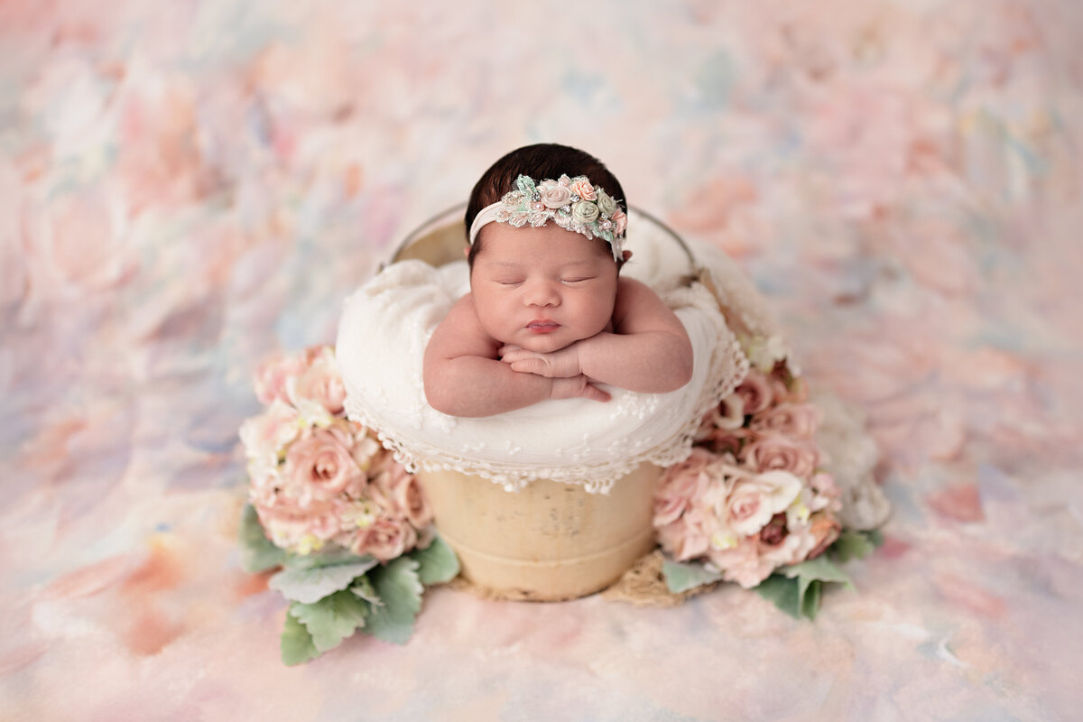 Newborn in a peach-floral wreath bucket posed on a pastel backdrop