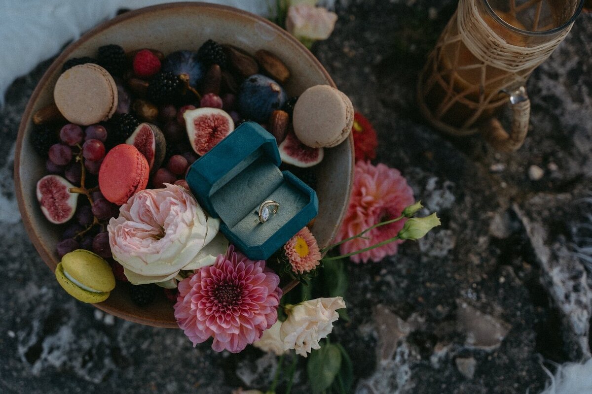 Bague de fiançailles dans son écrin cachée au milieu d'une corbeille remplie de fruits, de macarons te de fleurs. Séance photo engagement par Kamescop, vidéaste et photographe vintage en bretagne.