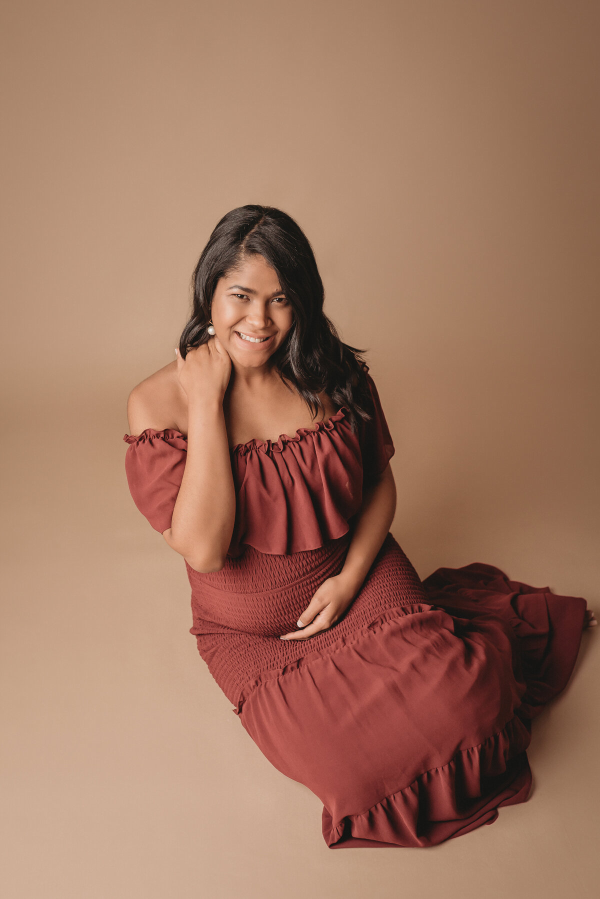 Pregnant woman at 36 weeks wearing burgundy dress posing for maternity portraits on a tan backdrop.