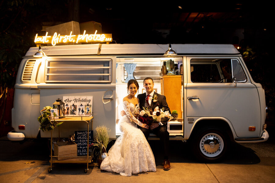 A bride and groom sit in front of a vintage VW bus turned into a stylish wedding photo booth, illuminated by warm neon lights for a fun and unique wedding experience.