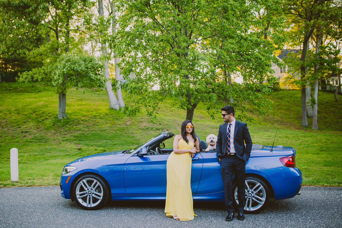 A couple standing in front of their blue bmw convertible with the dog popping out of the backseat at callahans beach, long island ny