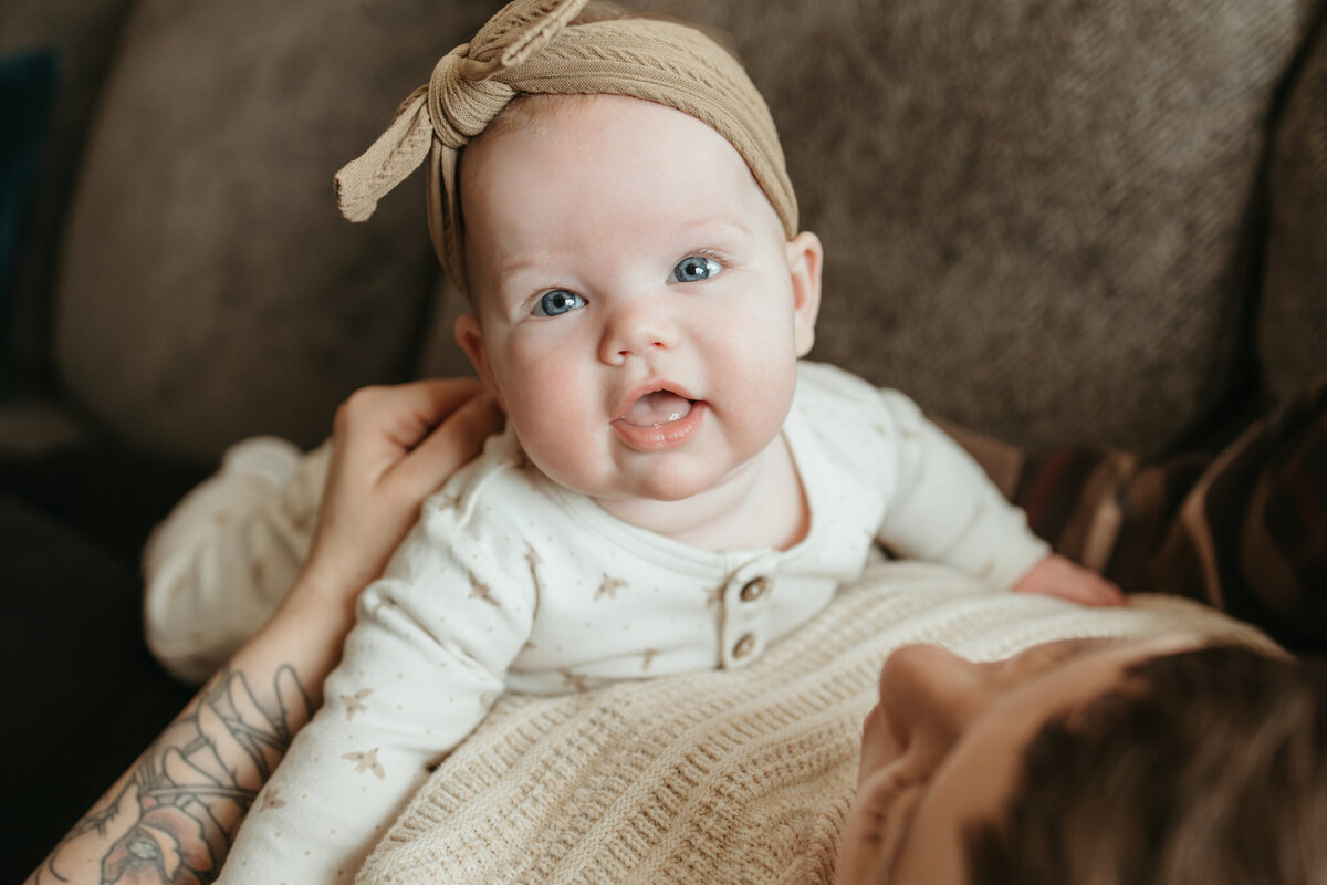 baby girl with tan bow and white outfit smiling and looking at camera during in home family session with alexis adkins photography in longmont colorado