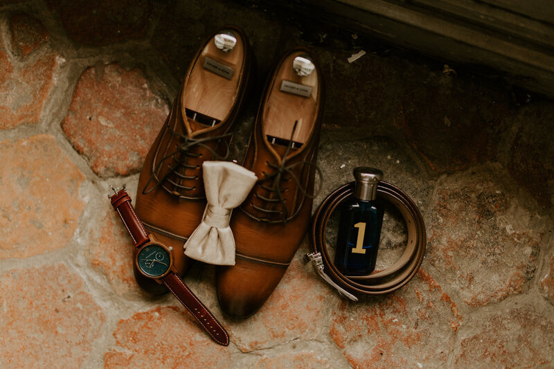 Chaussures de marié posés sur des tommettes avec une montre, un noeud papillon, une ceinture et un parfum. Composition par Laura, photographe de mariage en vendée.