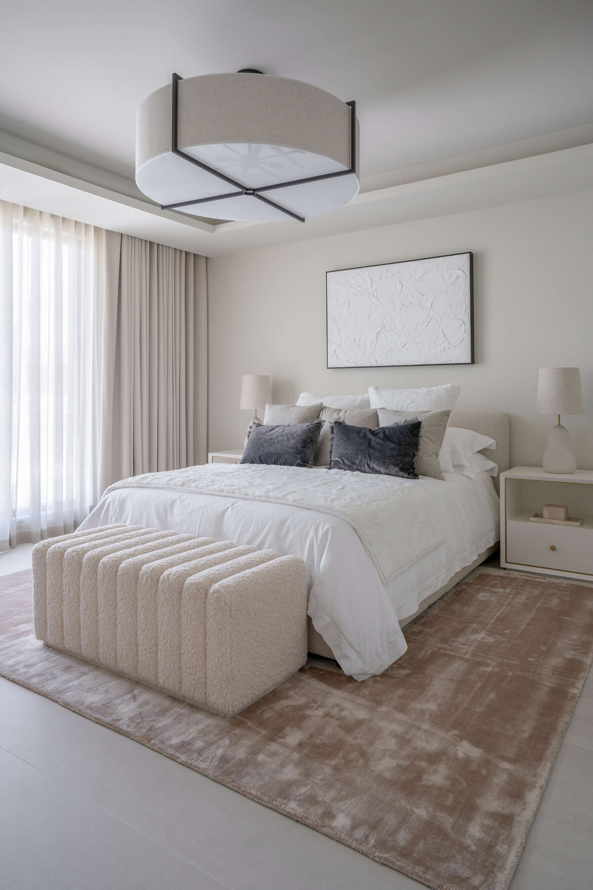 Light and airy guest room featuring a white bed, cream colored bench, and white side tables atop a soft tan rug. Above the bed is a white art piece. On the ceiling there is a large, round light. Floor-to-ceiling drapes cover large windows extending the length of one of the walls.