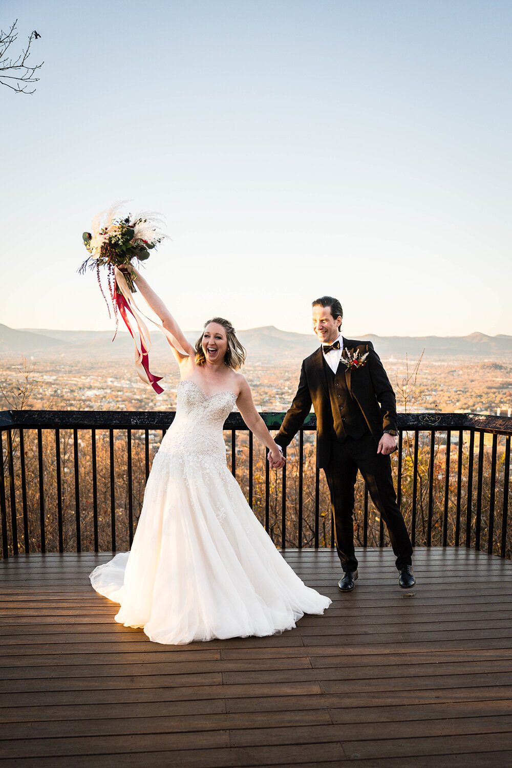 A newlywed couple holds hands on their elopement day for a portrait at the Rockledge Overlook at sunset in Roanoke, Virginia. The bride holds her bouquet in the air and has an excited expression on her face while the groom smiles widely.