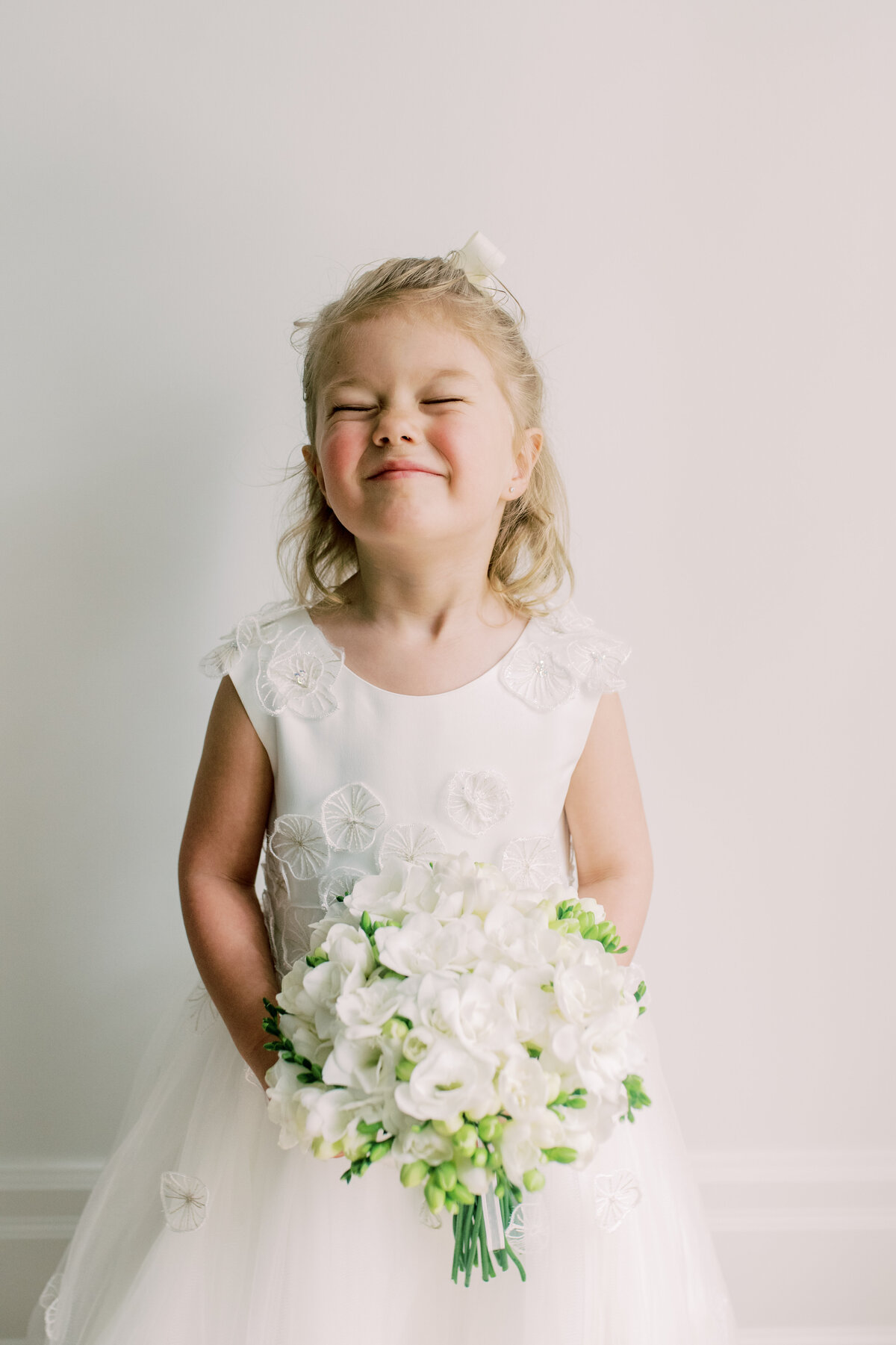 An adorable close-up of a flower girl in a white dress, beaming with a playful, scrunched smile while holding a bouquet of white flowers