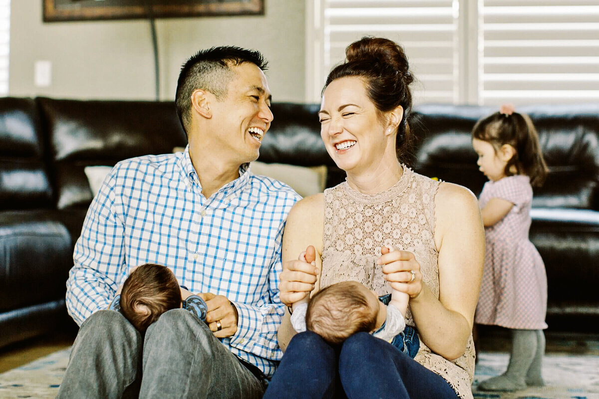 parents laugh while holding twins and sitting in front of toddle girl in their living room