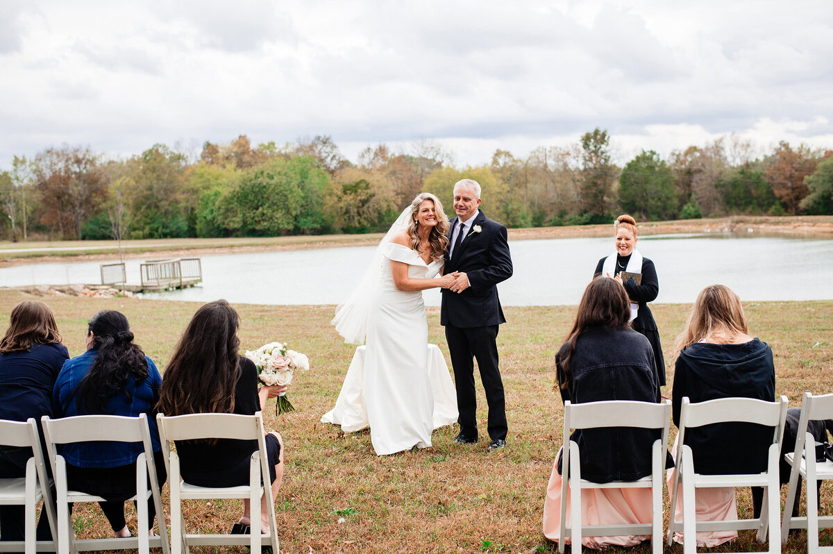 Bride and Groom holding hands and laughing together during their vows at Sugarfoot in front of a pond