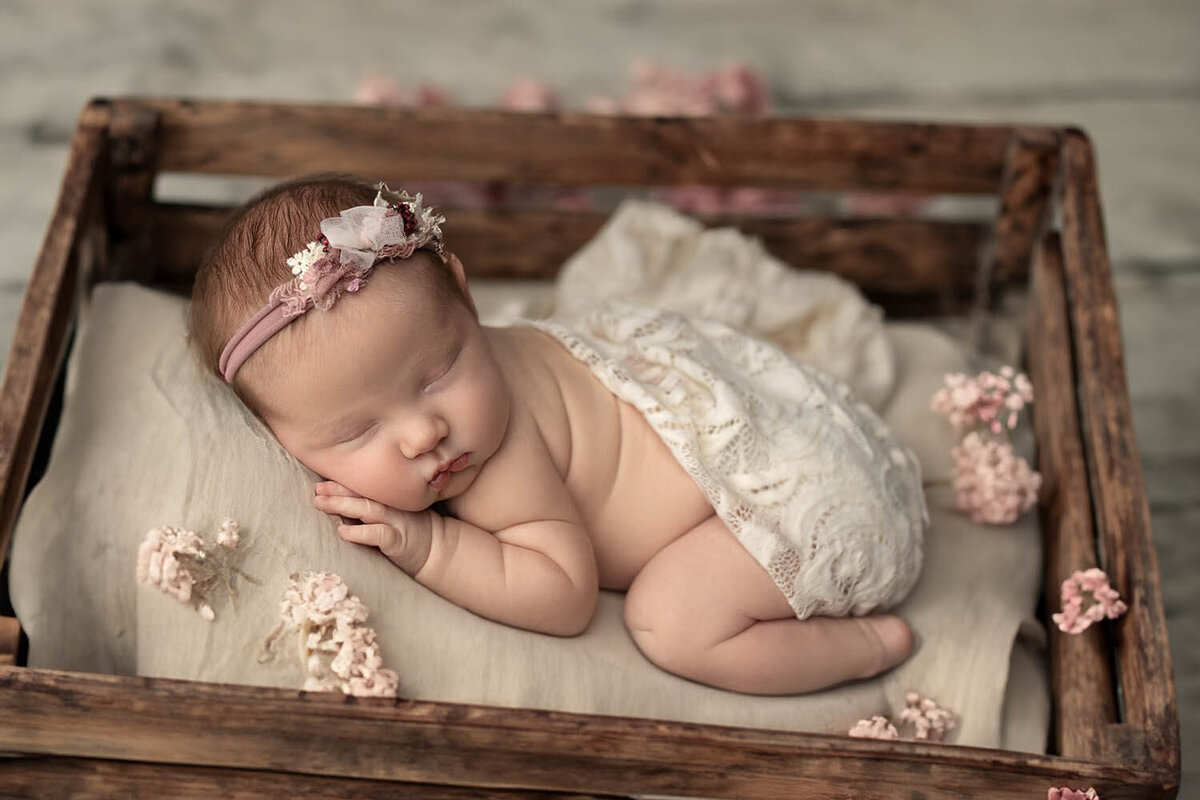 A newborn baby sleeps in froggy pose inside a wooden crate with pink flowers
