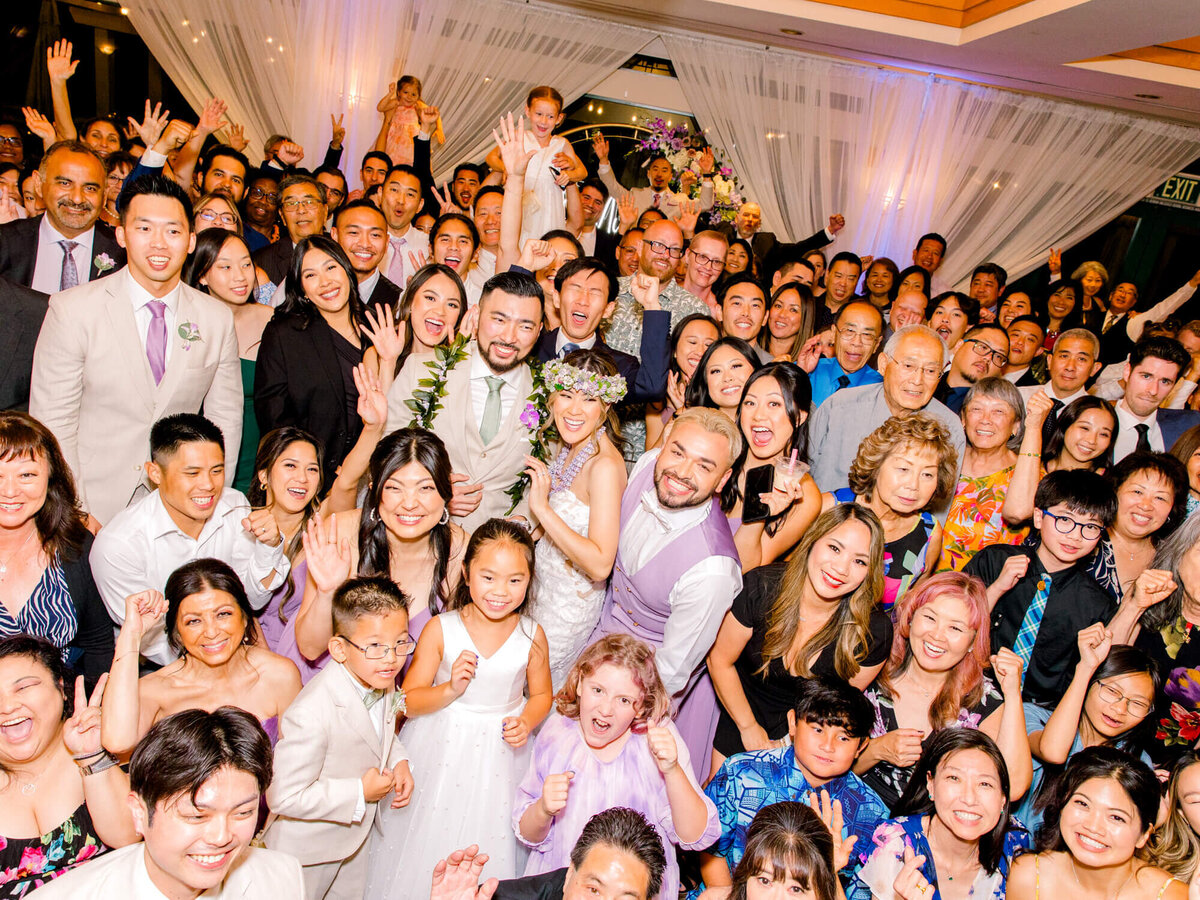 A joyful, diverse wedding group celebrates, cheering with smiles and raised hands. The couple stands centered under soft drapery, exuding happiness.