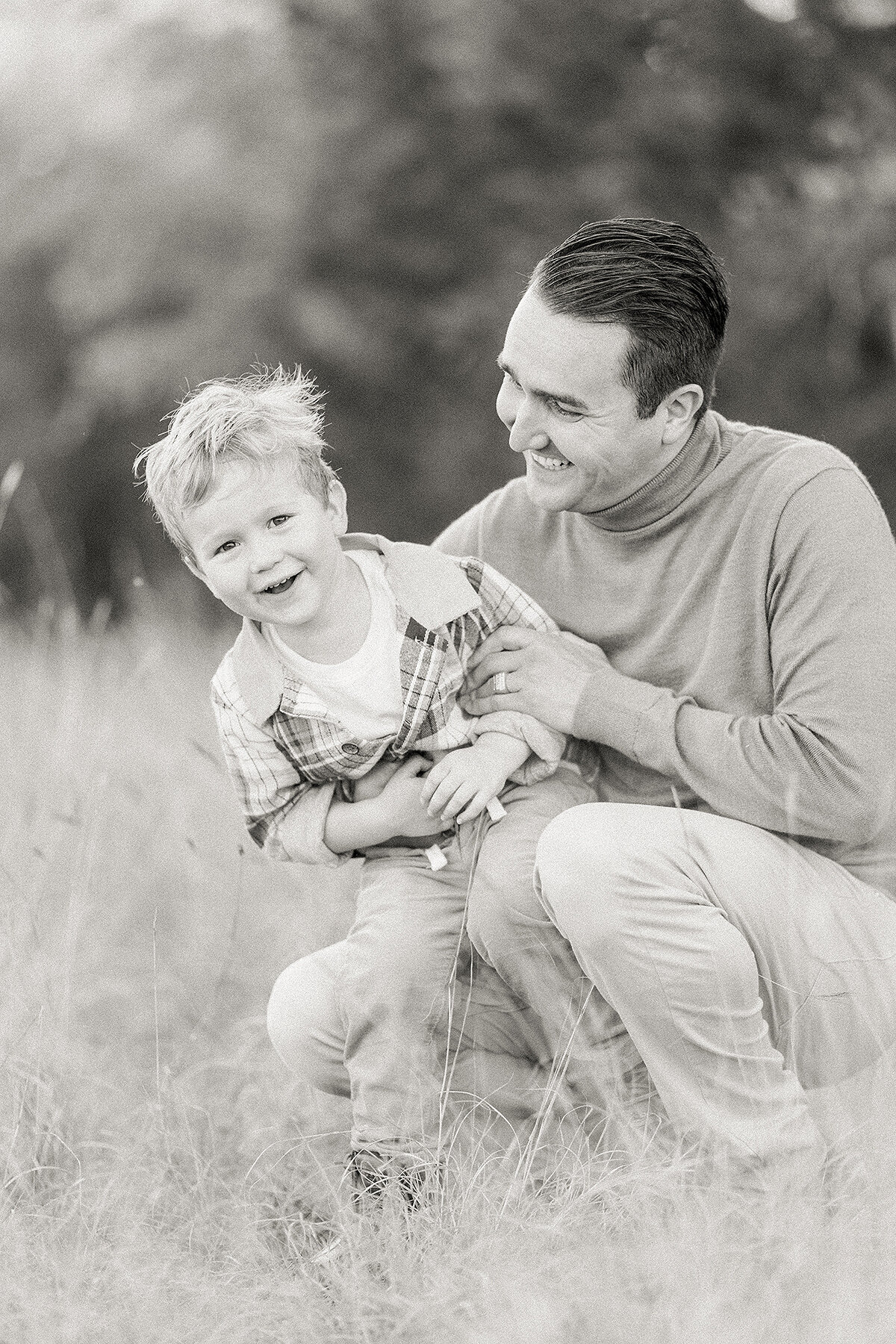 Black and white photo of a dad kneeling down in the grass while he holds his son for family photos.