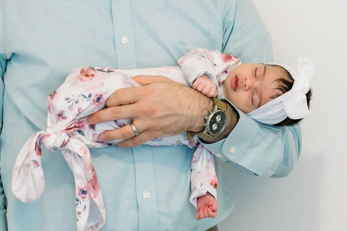 A sweet father-daughter moment captured by a Birmingham newborn photographer. A tiny newborn baby girl sleeps peacefully in her dad’s arms, wrapped in a floral knotted gown with a matching bow. The soft, neutral backdrop highlights the tenderness of this in-home lifestyle newborn session.