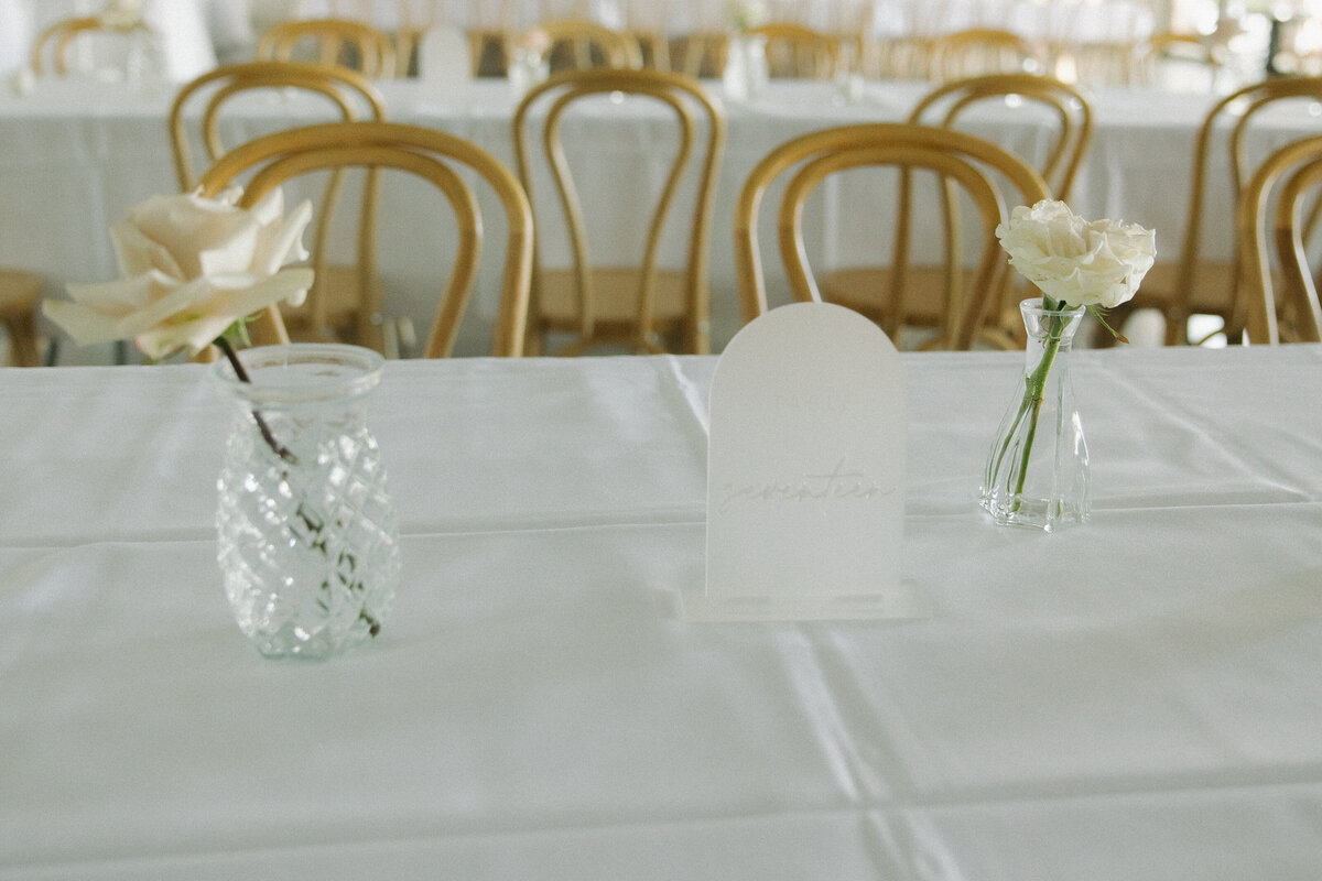 Two bud vases with a white opaque table marker. There are light pink roses in each bud vase.  There are light wood chairs behind the bud vase.