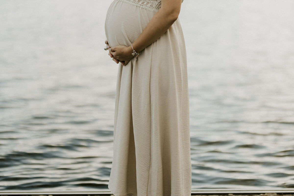 Femme enceinte de profil, en robe longue fluide pose tenant son ventre devant un lac pour Laura, lors d'une séance photo grossesse en vendée.