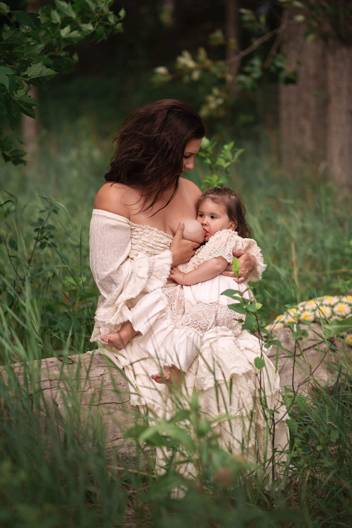A beautiful mother sits on a log and breastfeeds her infant daughter. Moms looks at daughter. Brittany danielle photography