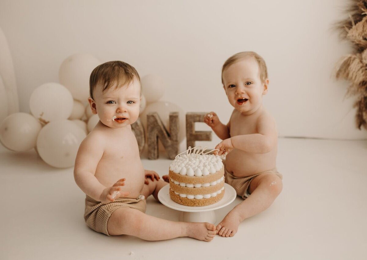 Twin babies enjoying a shared cake smash moment with matching outfits and a rustic “ONE” sign in background.