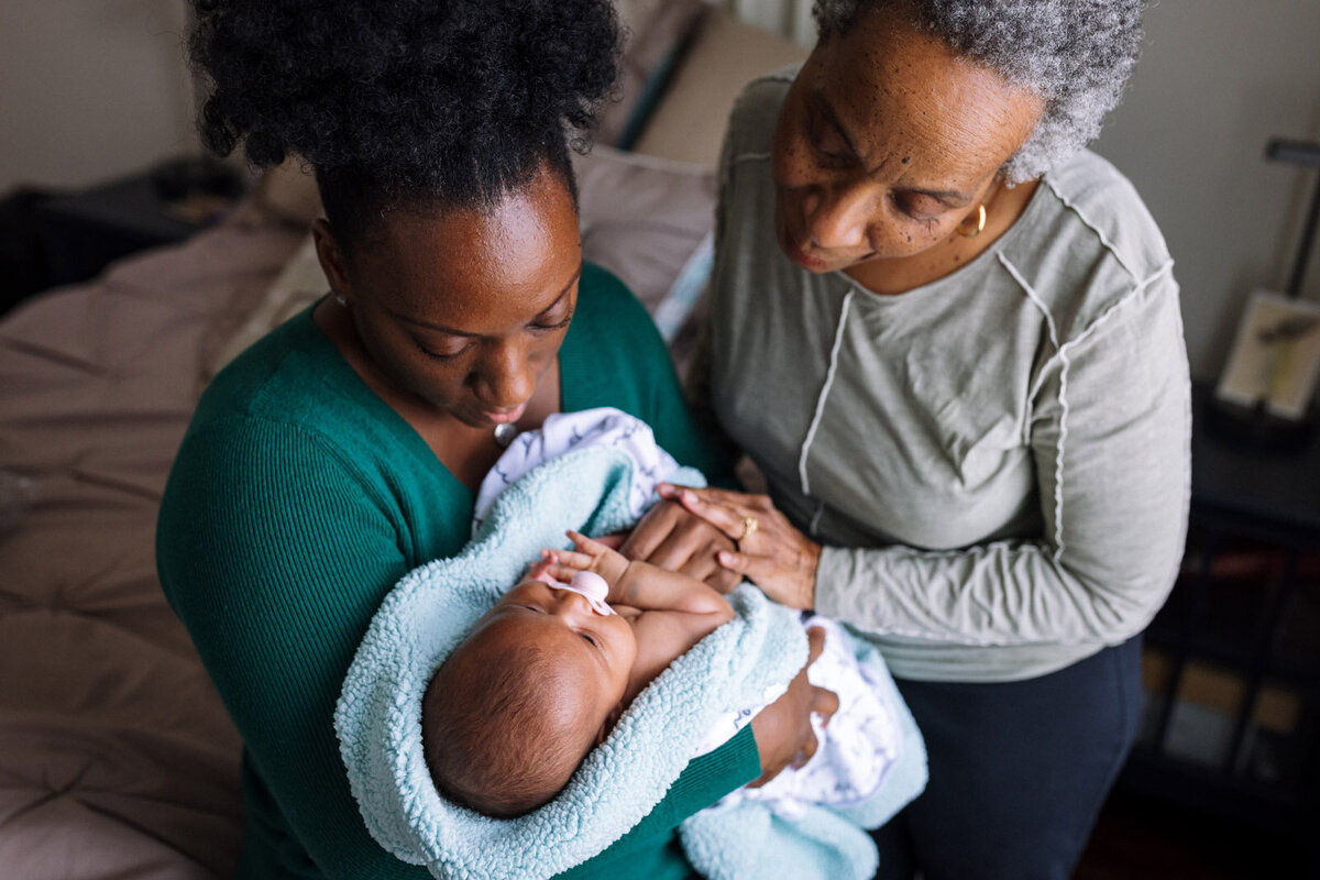 mom holding newborn wrapped in light blue blanket with grandma touching mom's hand and looking at baby