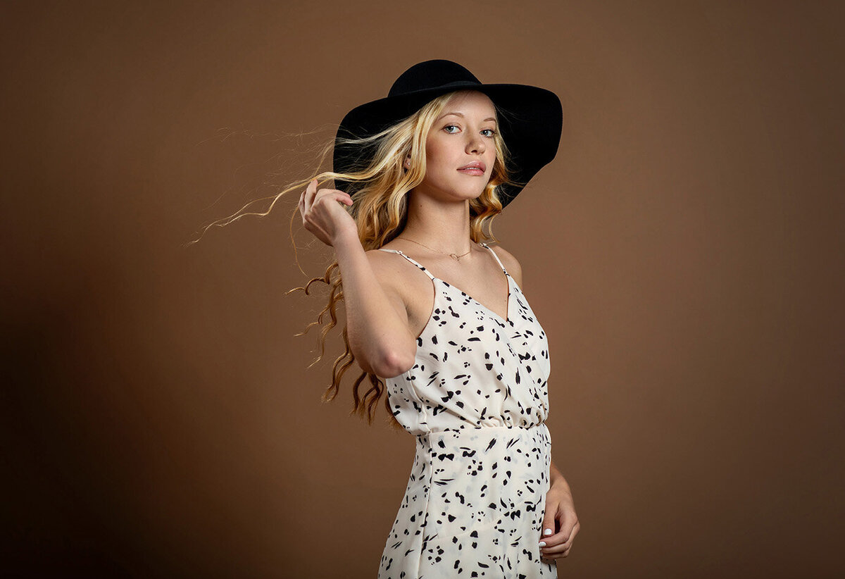 High School senior studio portrait of tall blonde girl wearing black hat and white dress with fan blowing hair.