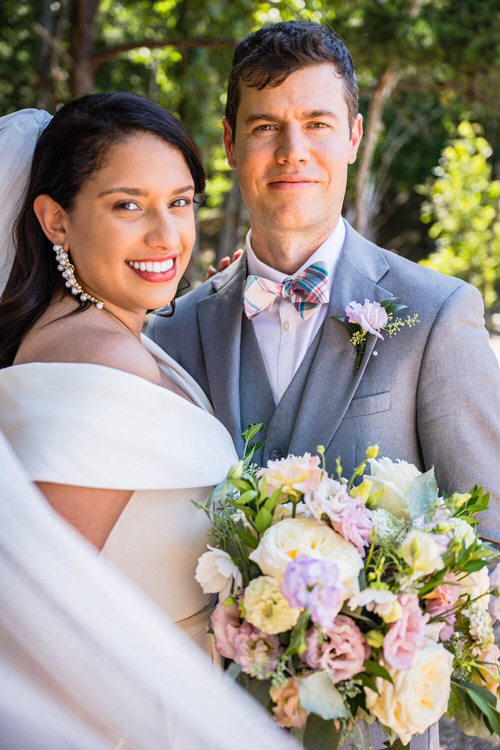 A bride and groom take a formal portrait together under the shade of some trees at Carvin’s Cove in Roanoke, Virginia. The groom is straight on while the bride is slightly angled. The bride’s bouquet takes up the majority of the bottom right of the photo while her veil curves ever so slightly towards the bottom right as well, so as to frame the individuals together.