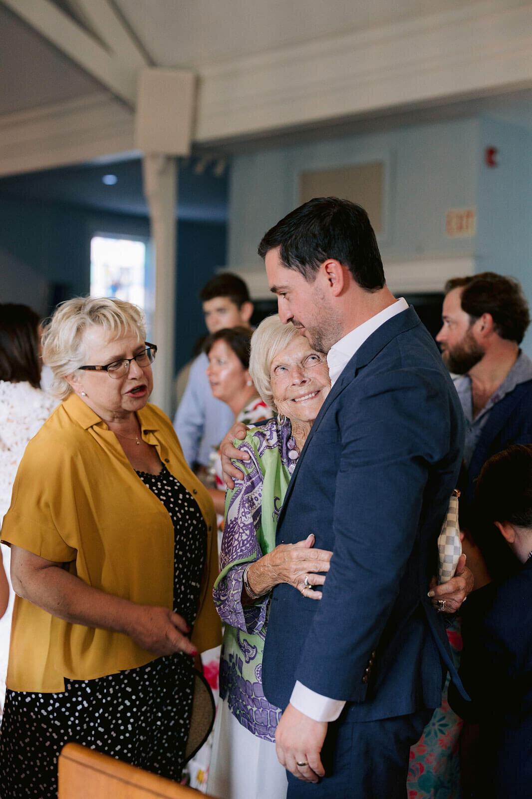 The groom is being hugged by an old woman while he speaks to another old woman at Cape Cod Summer Tent.