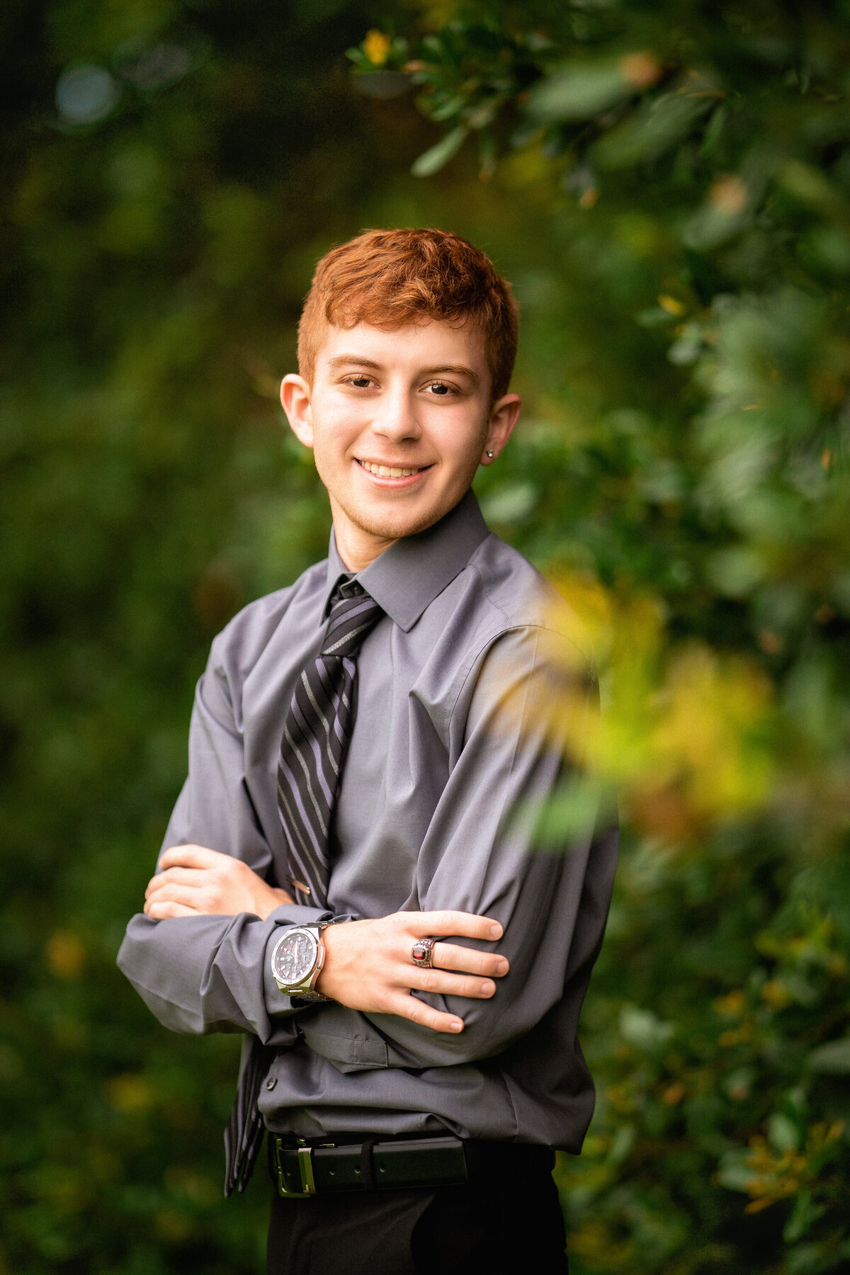 Senior boy with red hair standing in  the trees.  He has his arms crossed over his chest.  He is wearing a gray dress shirt and black striped tie.  He is also wearing a class ring and a watch.  He is turned slightly and facing the camera.