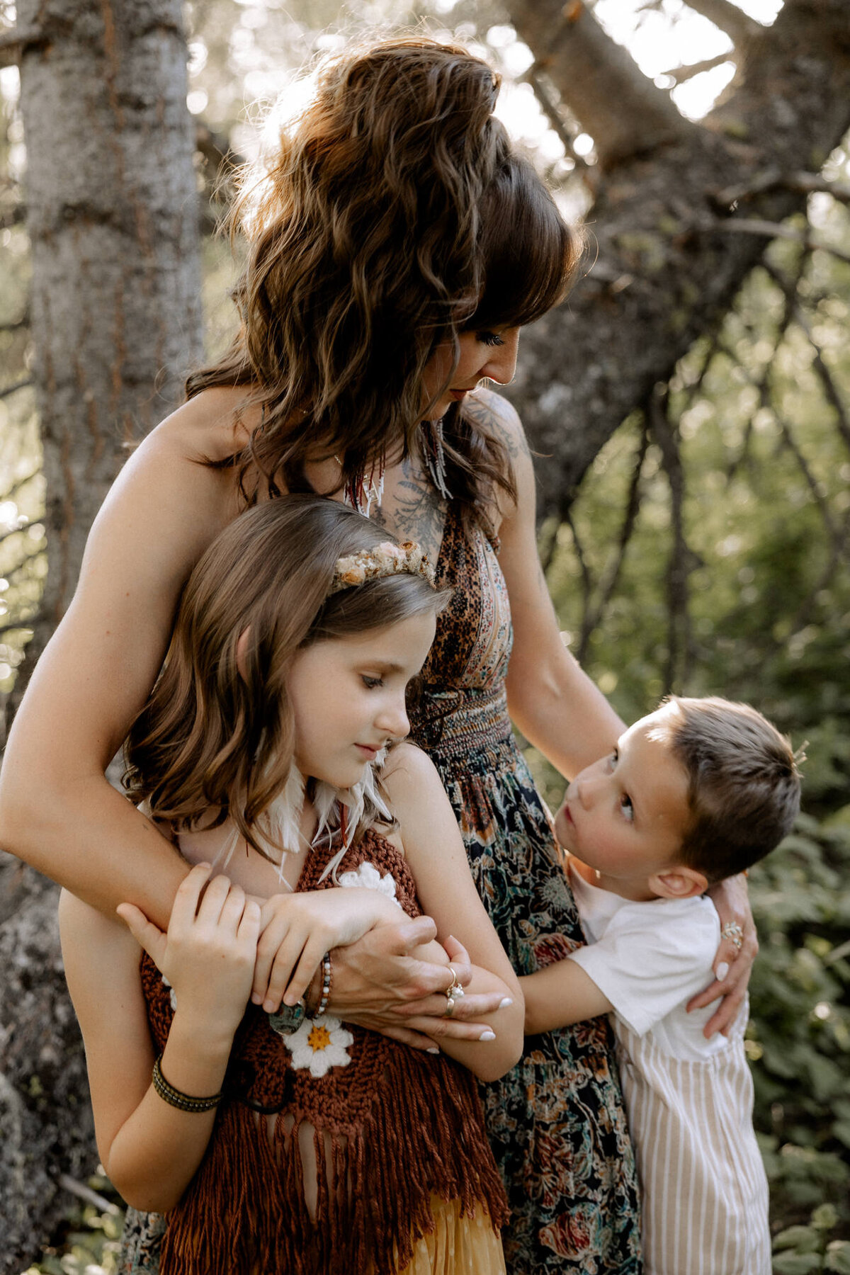 A tender motherly embrace in a woodland setting—Mom, dressed in a bohemian-style dress, wraps her arms around her daughter while her young son gazes up at her with love. Soft sunlight filters through the trees, highlighting this heartfelt family moment.