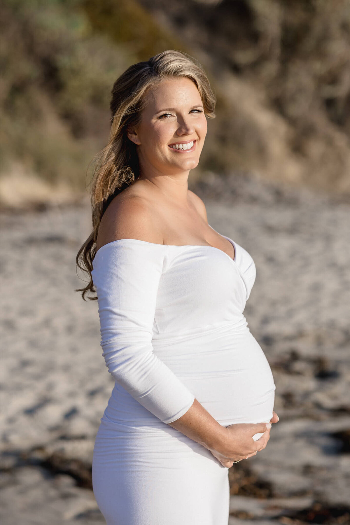 A smiling pregnant woman stands on a sandy beach, wearing a form-fitting, off-the-shoulder white dress. She gently cradles her baby bump with both hands. Driftwood and blurred bushes are in the background under natural light.
