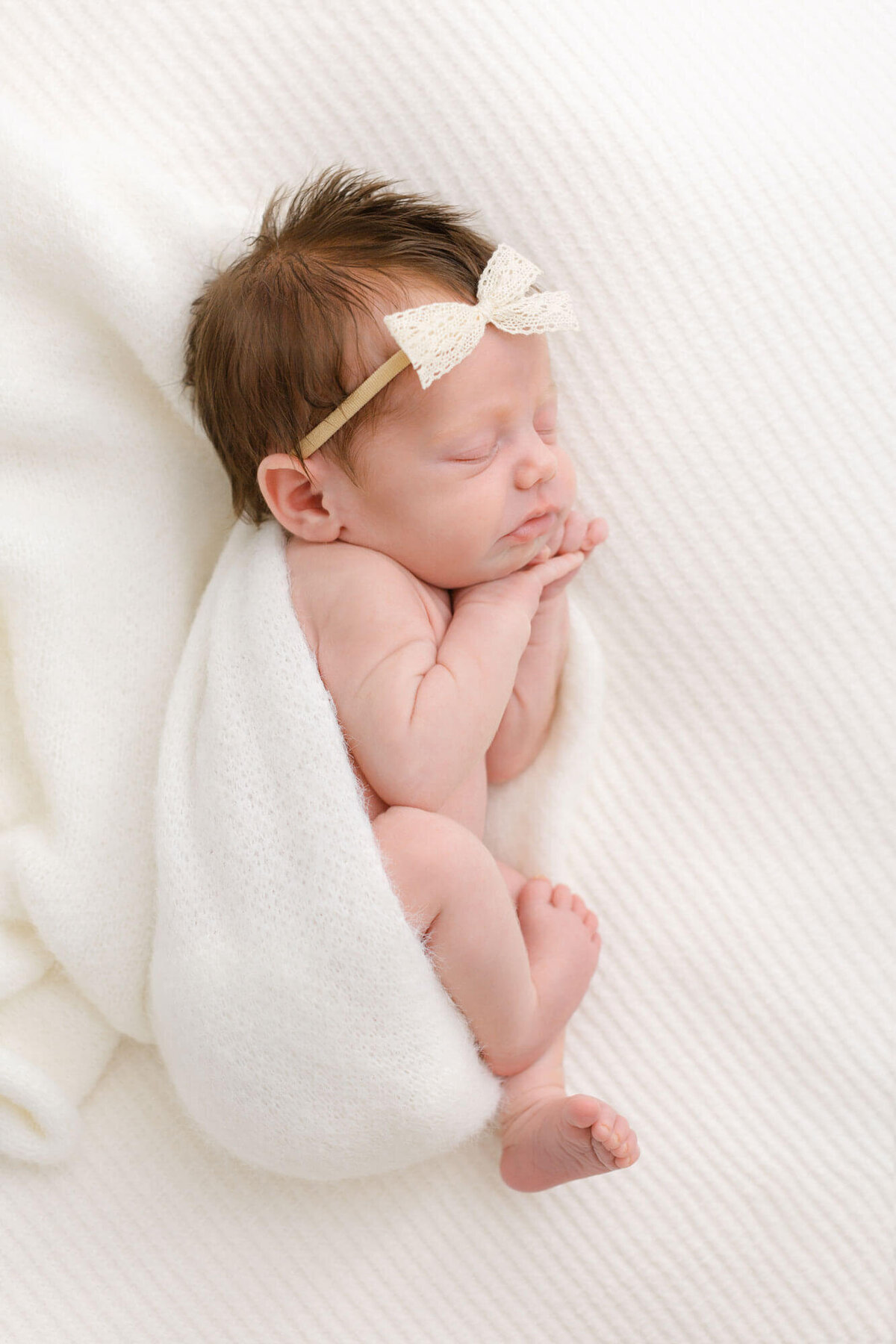 Newborn baby laying on side with a cozy soft white blanket wrapped around the back of her body. Her hands are up by her face and her feet are hanging out a bit and she has a little white bow on her head.