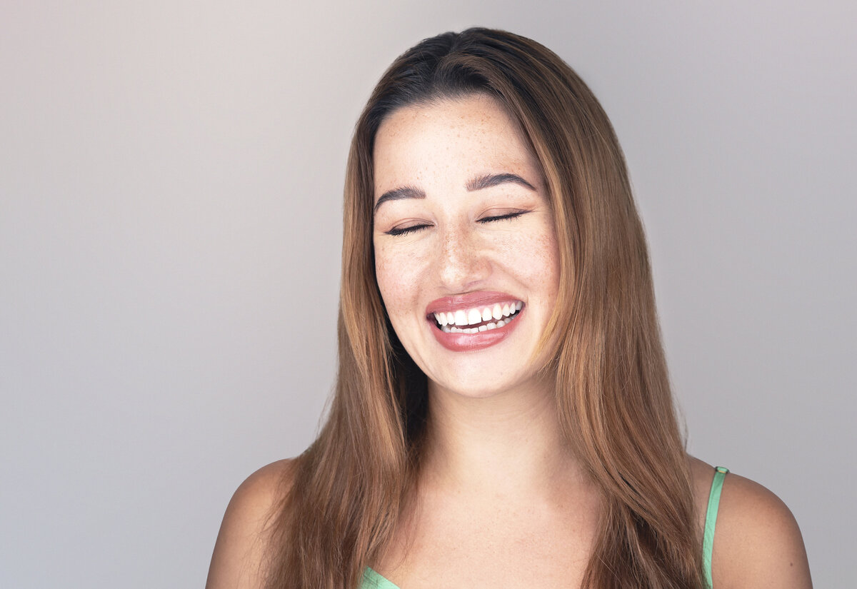 Woman in green tank top with long brown hair poses for a studio headshot. Her eyes are closed and she is laughing. Philadelphia and Main Line Headshot Photographer.