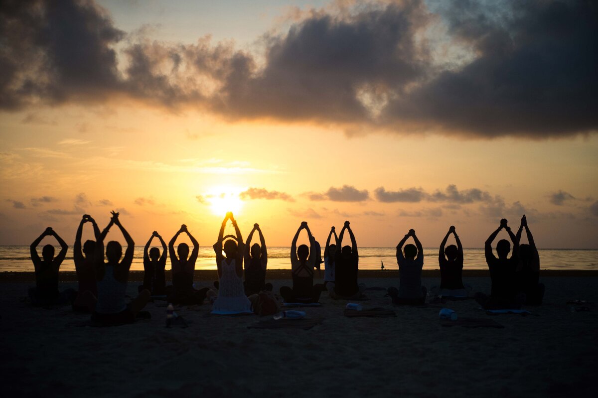 A group takes a Yoga class on the beach to show the tranquil and serene  setting. Participants have hands stretched out overhead silhouetted against sunset and sky.