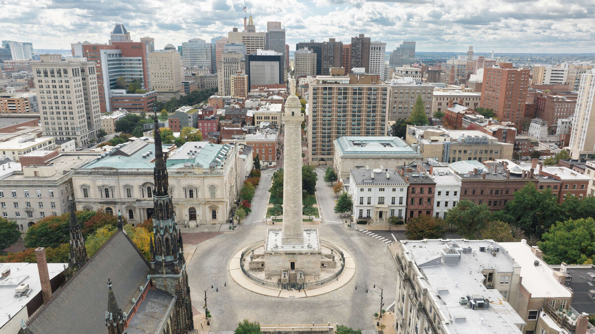 Aerial view of a cityscape featuring a tall, cylindrical monument surrounded by historic buildings and lush greenery. Skyscrapers rise in the background under a partly cloudy sky. The streets are lined with trees and cars.