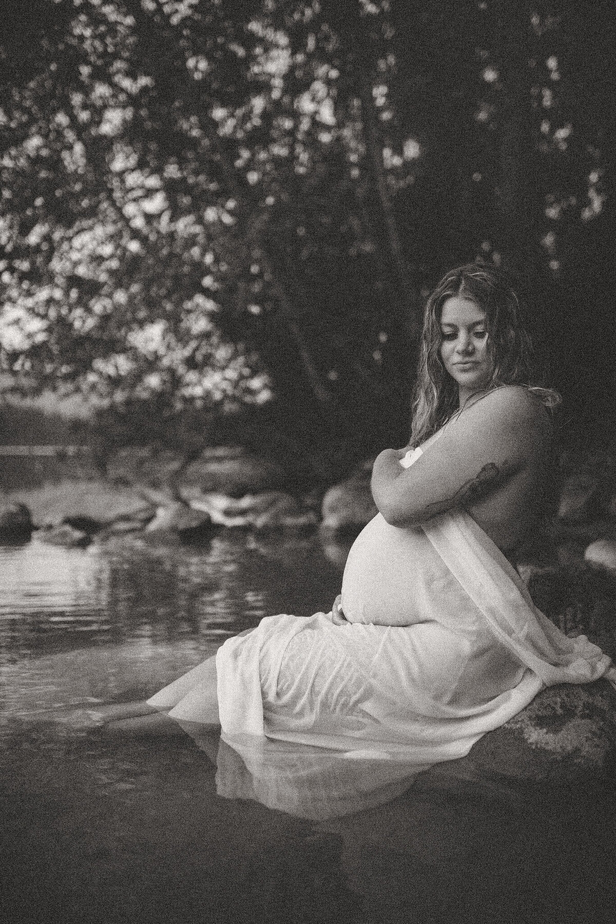 Brittany Danielle captures a black and white portrait of a pregnant woman sitting in water with a sheer white piece of fabric draped over her.