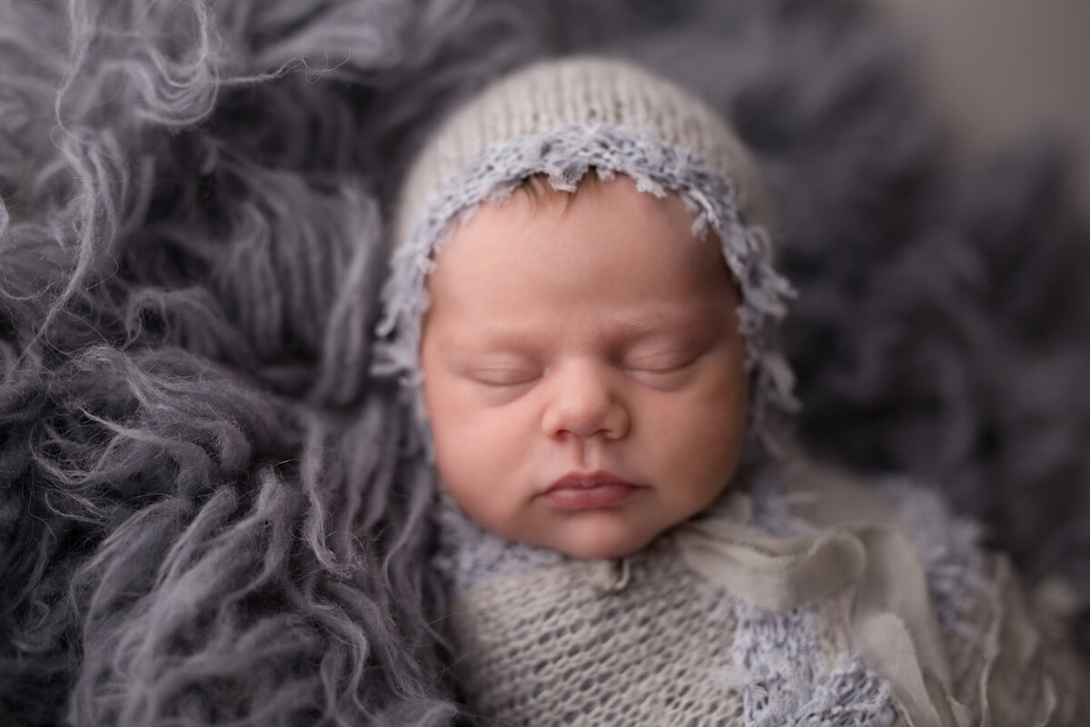 Baby girl in a bonnet and wrap on a gray fur backdrop.  She is asleep and her head is turned to the side.