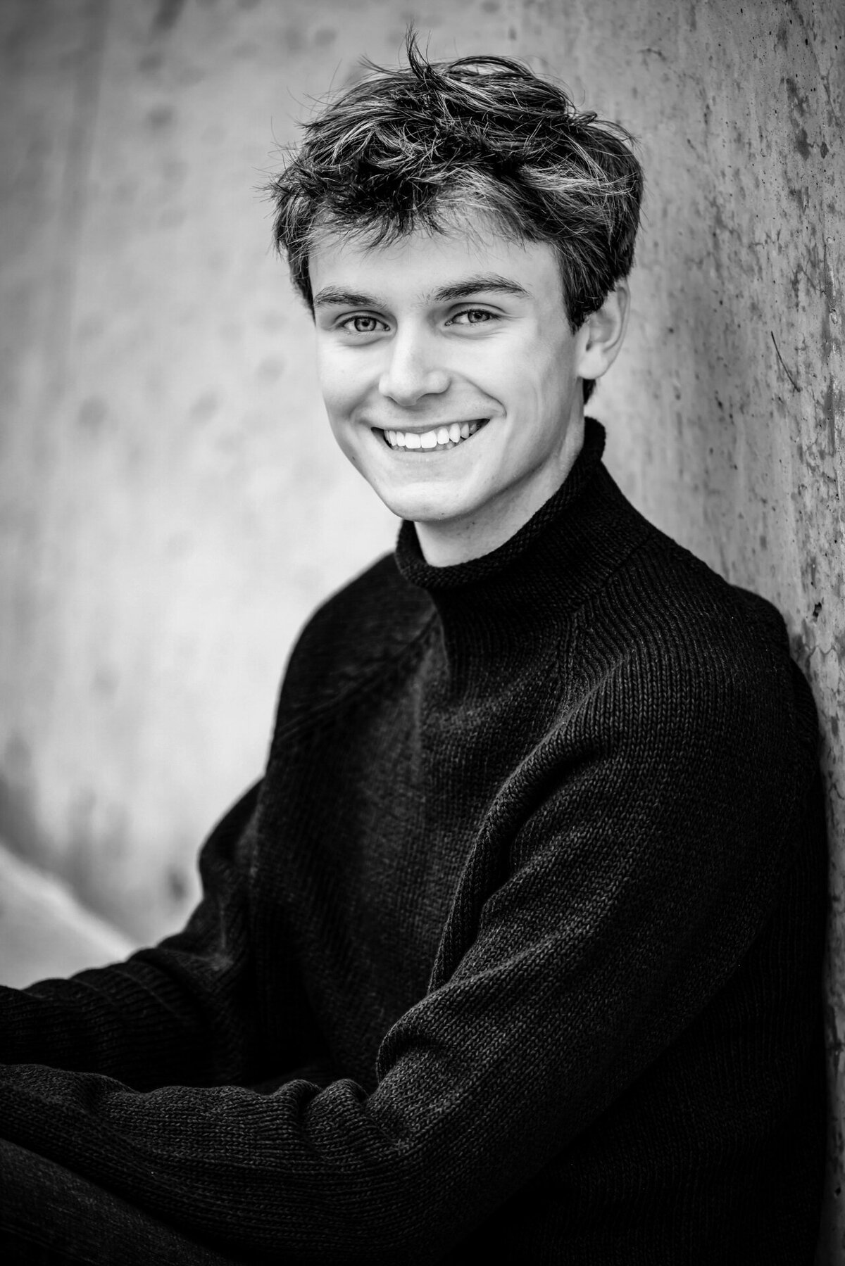 Black and white portrait of a senior boy in a black sweater, smiling naturally.