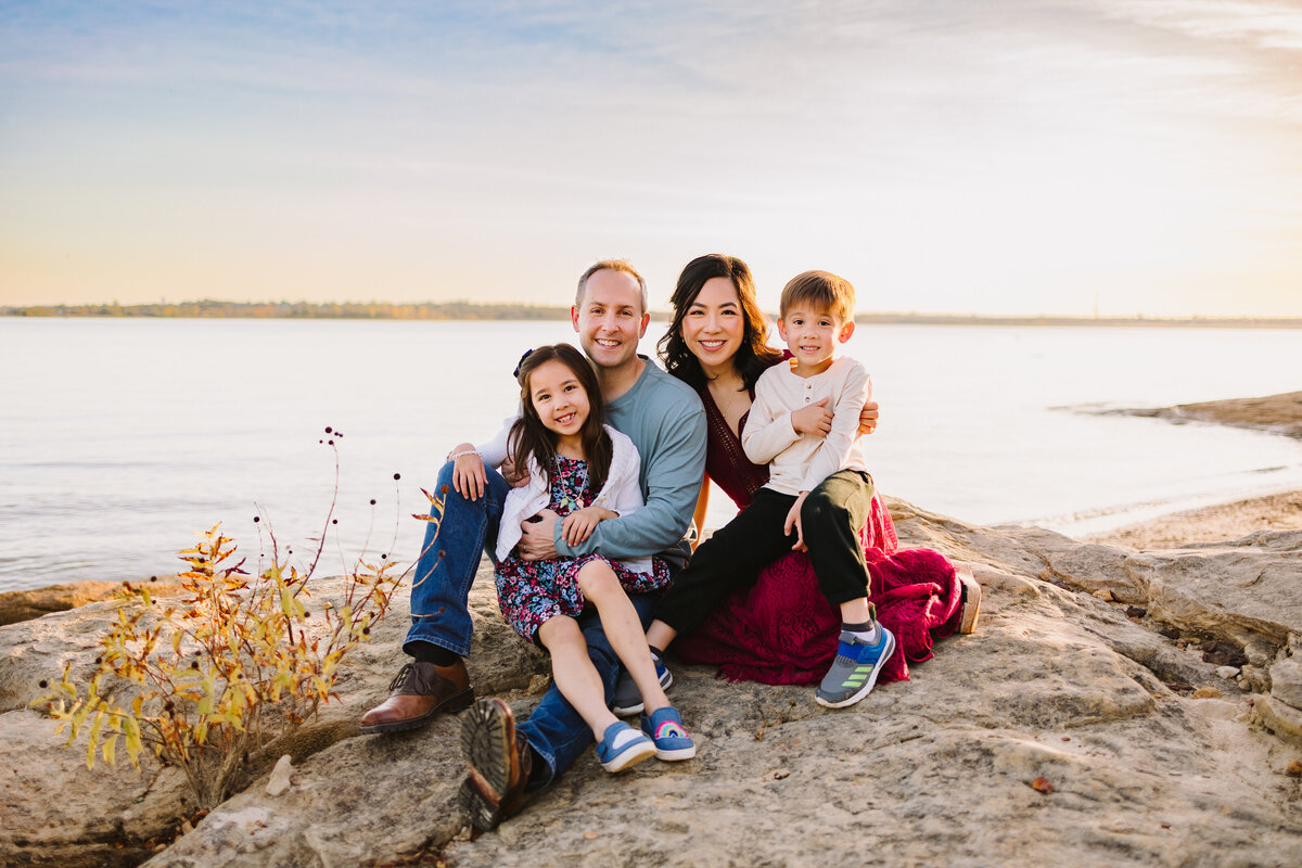 Family photography ideas in Albuquerque captured a memorable moment by a lake. The woman in a long red dress and white T-shirt is carrying a boy, while the man in jeans and a green T-shirt is holding a smiling girl dressed in a floral dress. The family is seated in front of rocks and a lake, all looking towards the camera.