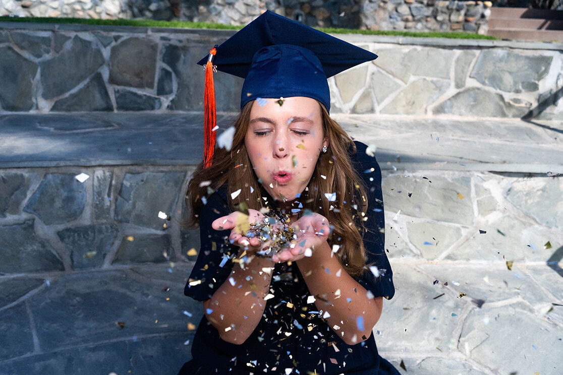 A graduate in navy and orange regalia closes her eyes, as she sits on large stone steps, and blows confetti into the camera from hands cupped to her mouth. Photo by SAVI Photography - Photographer in Riverside
