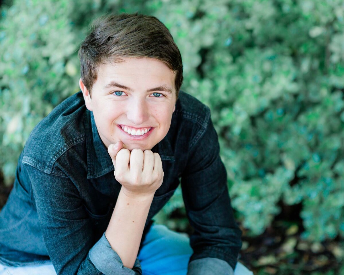 Senior portrait of a guy sitting on a bench leaning on his knees with green shrubbery behind him.