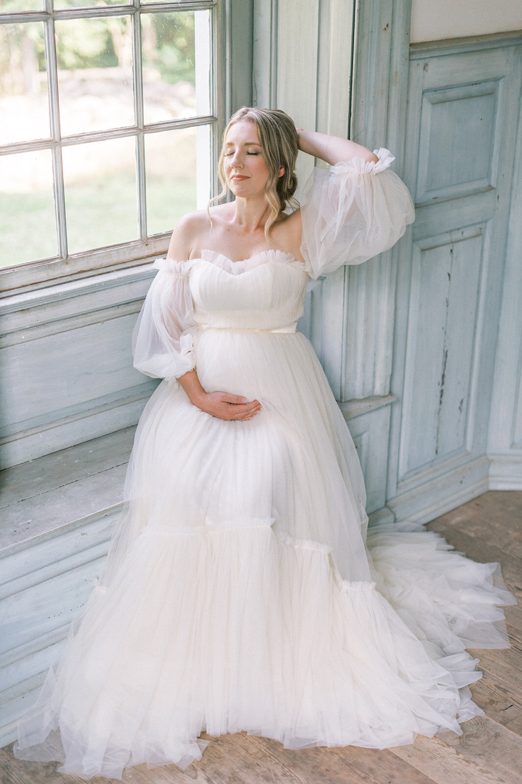 Pregnant mother sits in a window sill while wearing a flowy white gown during her maternity session at Salubria