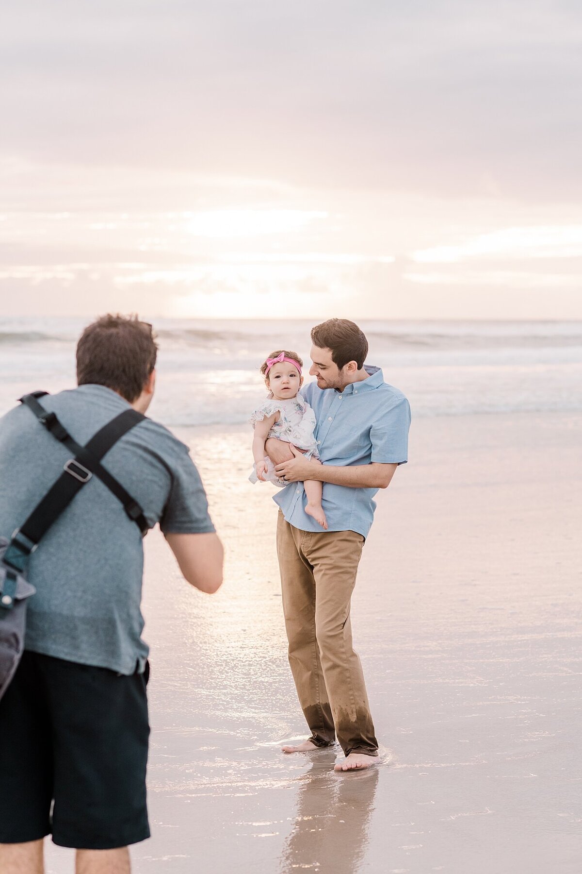 A father in a blue shirt and khakis holds his daughter on a beach while a photographer in a blue shirt takes their photo
