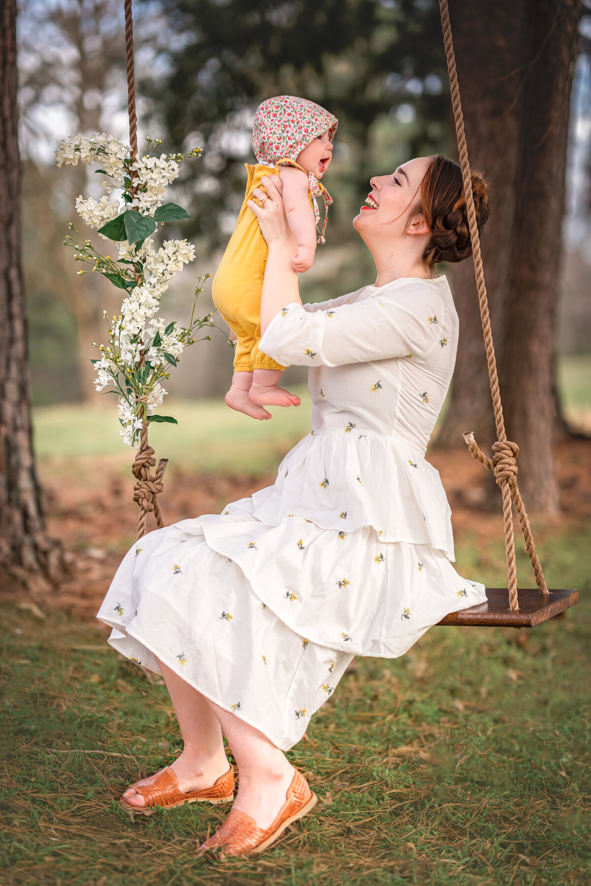 Mom in white dress sits in a wooden swing while she holds her baby.