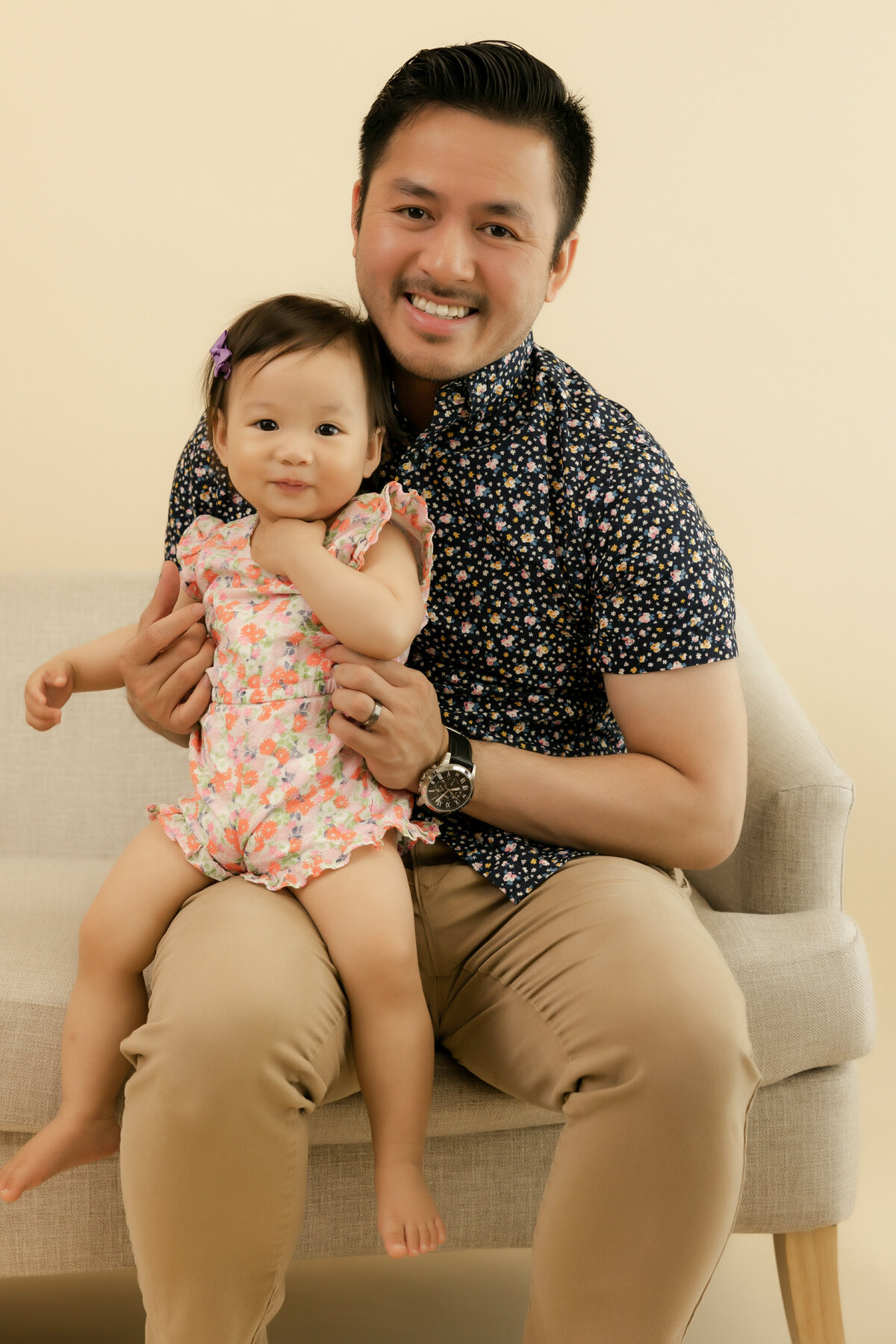 Dad sits on a beige couch holding his daughter on his lap smiling at the camera
