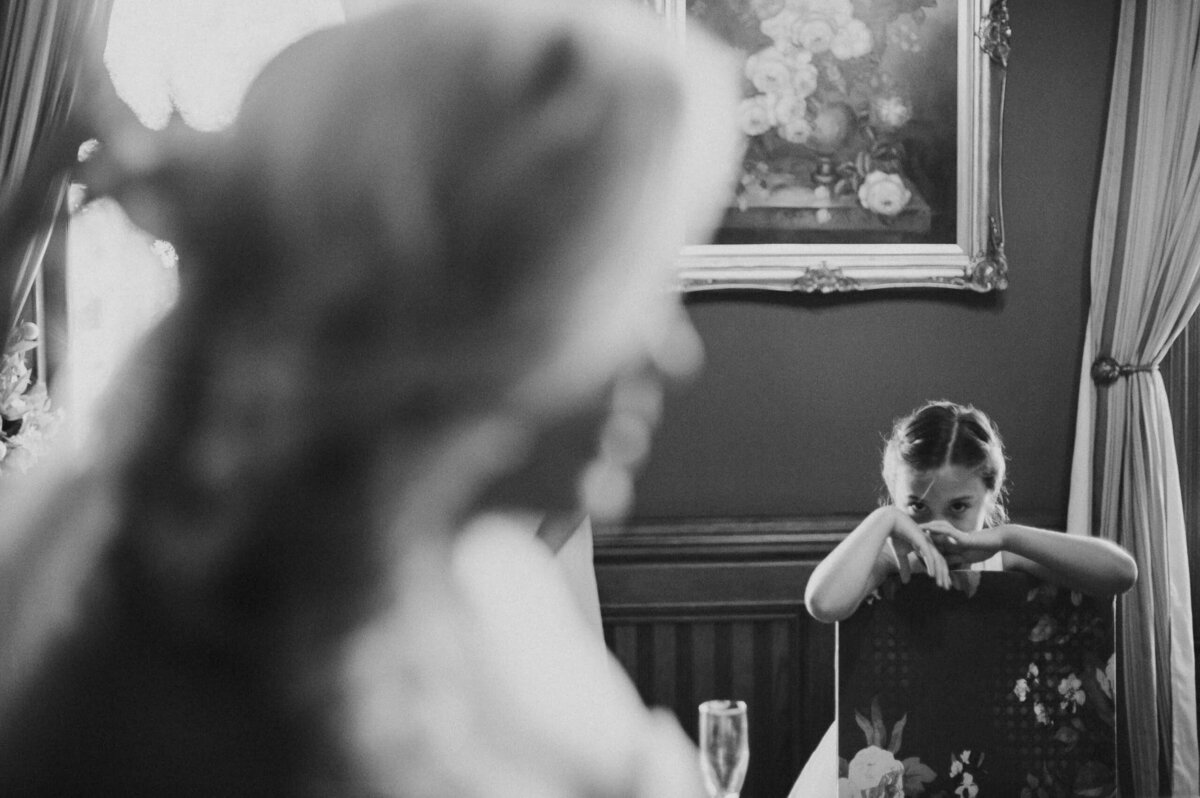A black and white image capturing the emotion of a young wedding guest covering her face with her hands, with the bride out-of-focus in the foreground