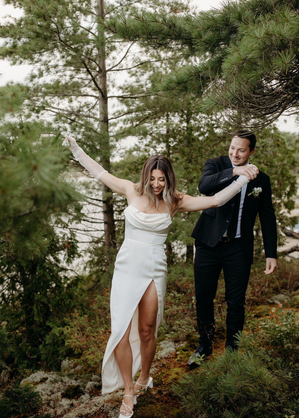 Bride in a chic white dress with a thigh-high slit and groom smiling as they navigate a rocky, wooded terrain