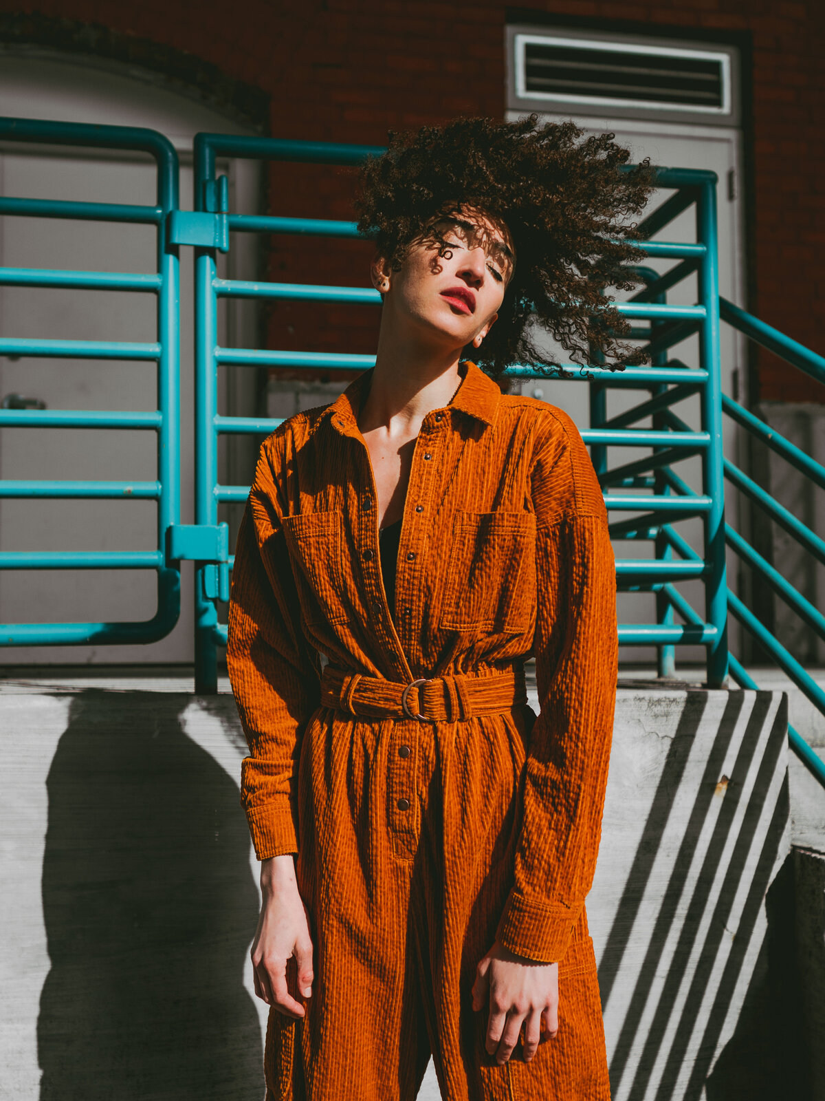 Woman in a textured rust jumpsuit standing against teal metal stairs, owning the sunlight – powerful fashion portrait by Mewphotovideo in Portland, Oregon