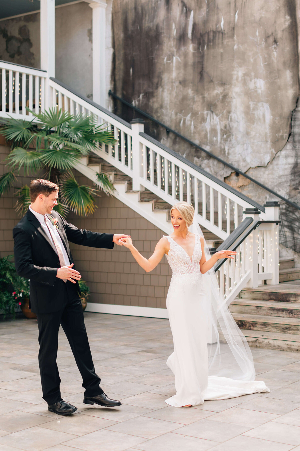 A bride and groom, both smiling, hold hands and dance in an outdoor courtyard. The bride wears a white gown and veil, while the groom is in a black suit. A staircase and green plants are in the background.