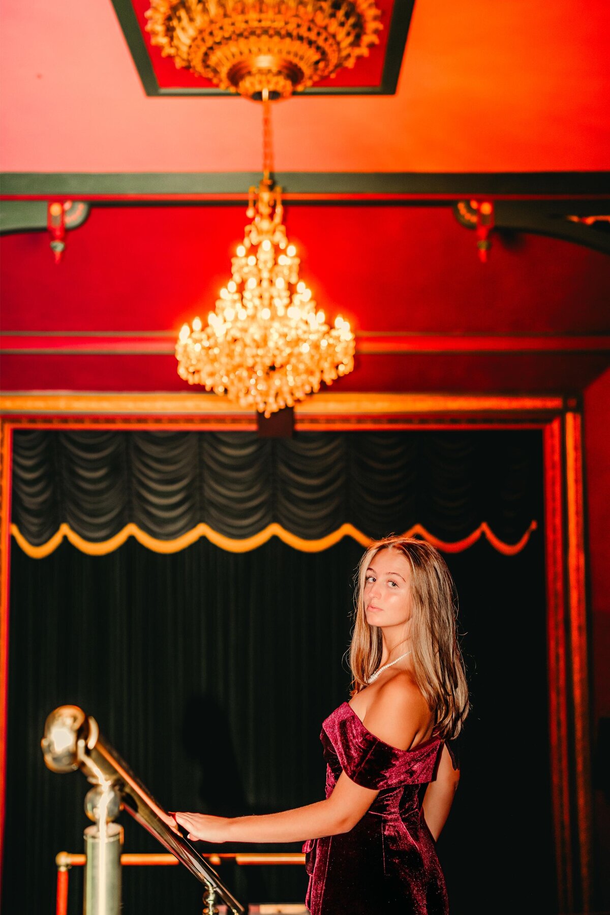 High school senior in a velvet dress stands under a glowing chandelier in a historic theater, capturing a moody & cinematic senior portrait.